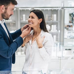 Beautiful couple enjoying in shopping at modern jewelry store. Young woman try it out gorgeous necklace and earrings.