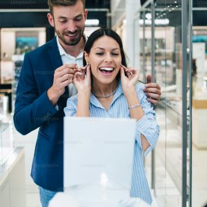 Beautiful couple enjoying in shopping at modern jewelry store. Young woman try it out gorgeous necklace and earrings.