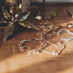 Modern gold and pearl accessories on table with vintage candlesticks. Stylish golden necklace and ring on wooden background in sunny light.